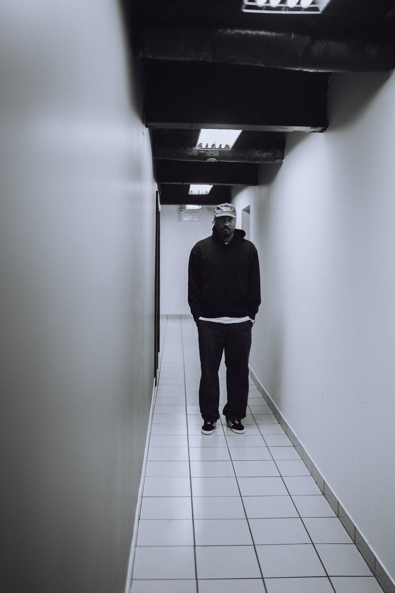 JEFFERY standing at the end of a white tiled hallway, hands in his pockets