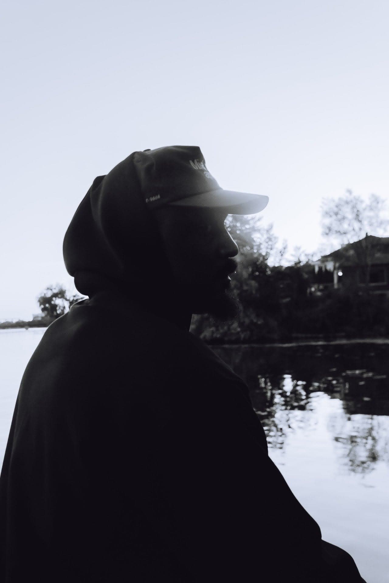 JEFFERY in silhouette by the water at dusk, hood up under his cap